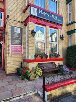 A brick building with a red awning and a sign that reads 'Happy Days Guest House.' There are flower pots on the sidewalk and a bench in front.