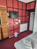 Habitación de hotel con diseño de panel de pared rojo y negro, un televisor montado en la pared, un radiador, un cubo de basura y una cama con manta a rayas.