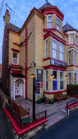Un edificio de varios pisos con fachada roja y beige, un letrero que dice 'Happy Days' y una banca en el exterior.