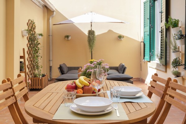 Een ronde houten tafel met een witte parasol, een schaal fruit en twee witte borden met bestek.