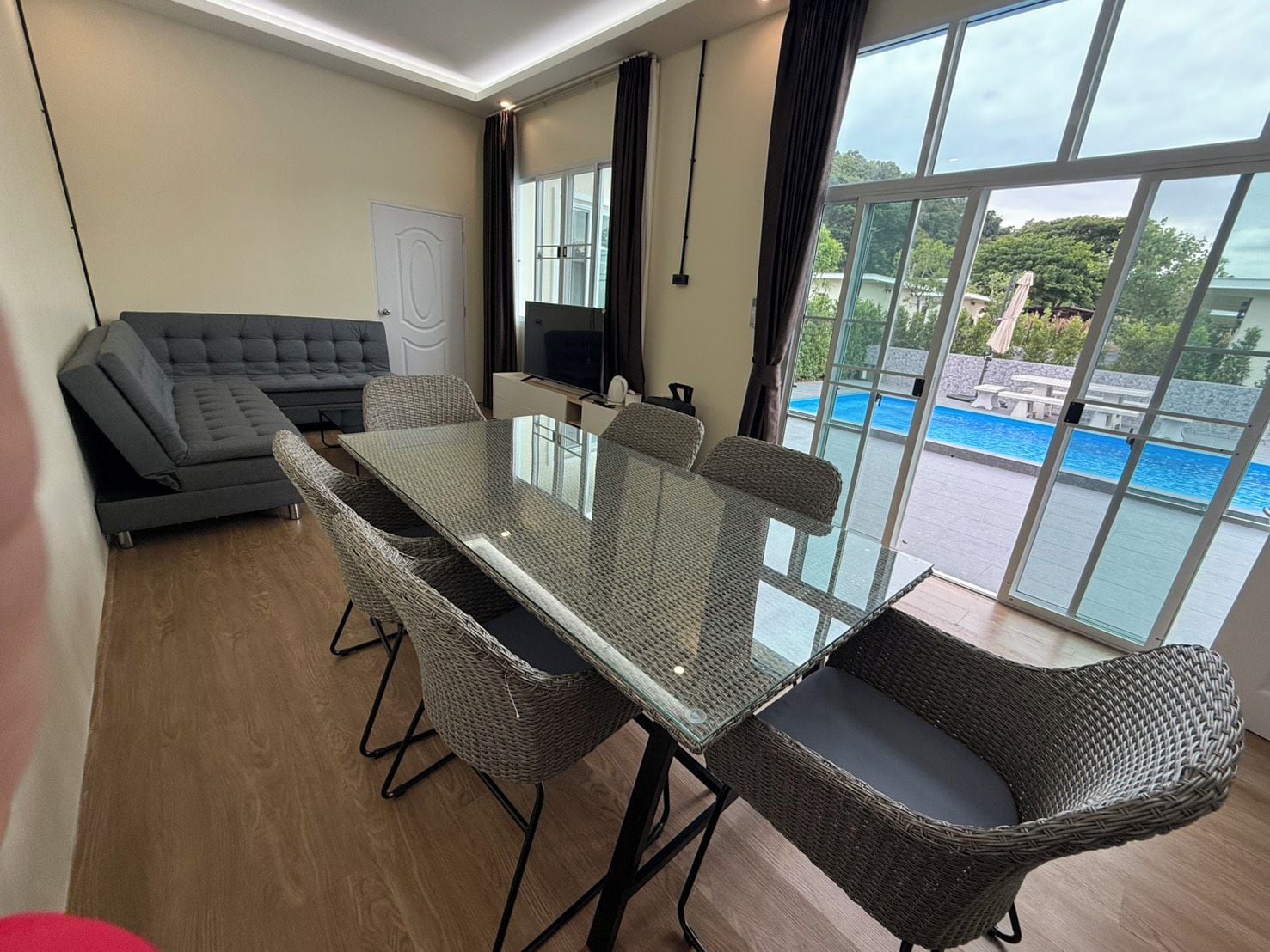 A room with a glass-top table, wicker chairs, a grey sofa, a flat-screen TV, and a view of a swimming pool.