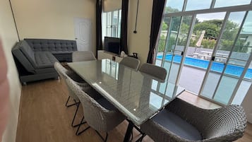 A room with a glass-top table, wicker chairs, a grey sofa, a flat-screen TV, and a view of a swimming pool.