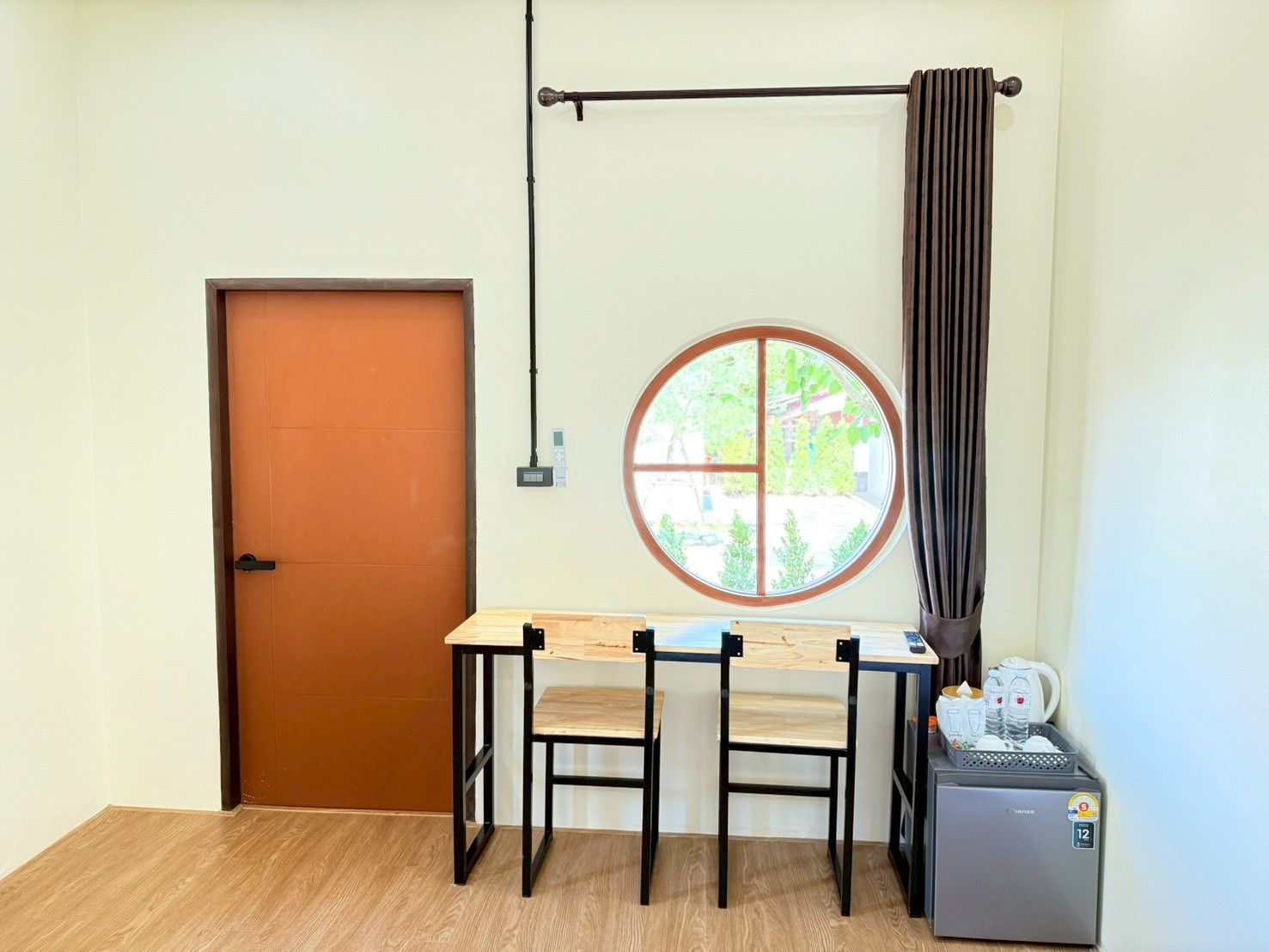 Quarto com porta de madeira, janela redonda com vista, mesa com cadeiras e um pequeno refrigerador.