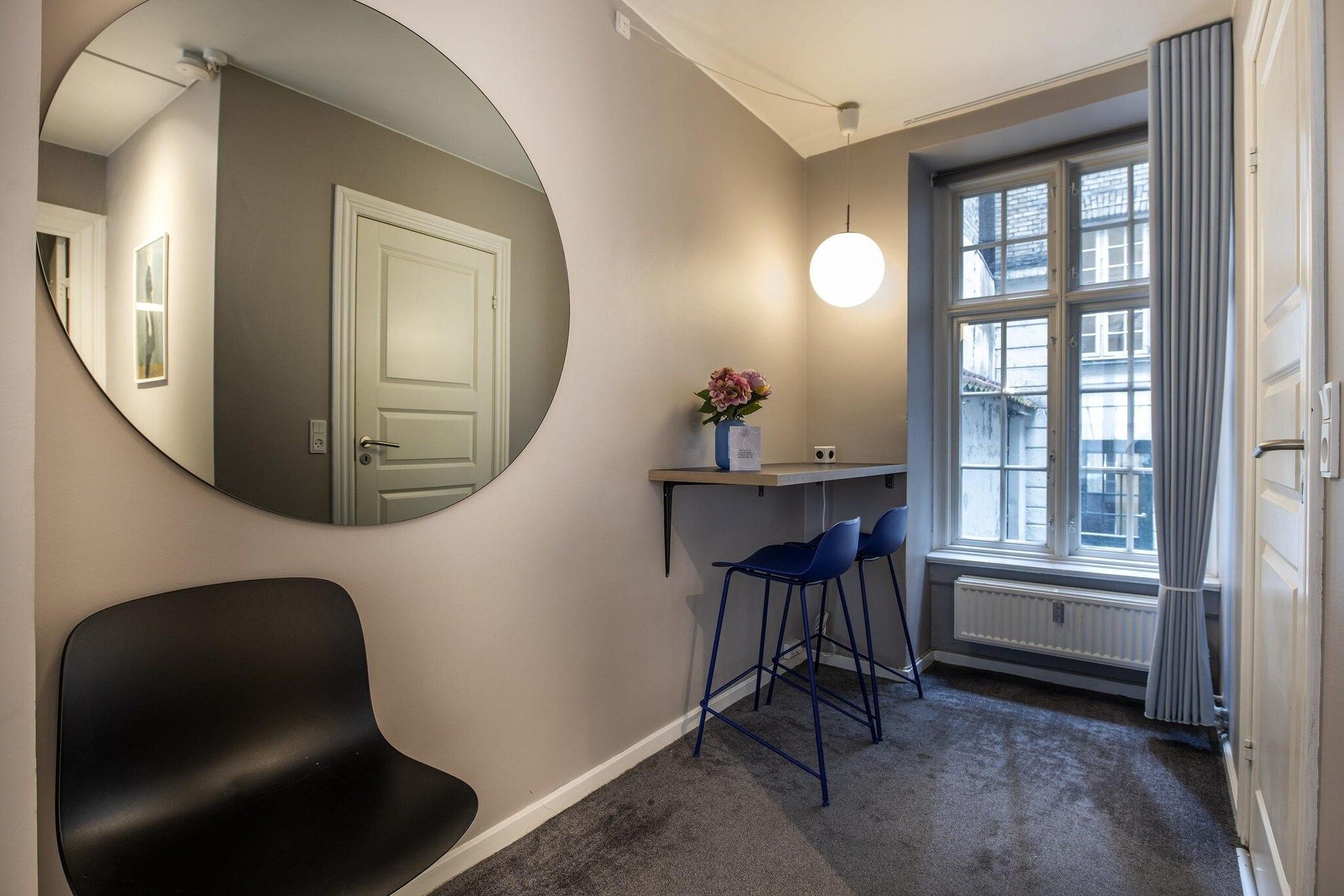 A room with a round mirror, a black chair, a desk with two blue chairs, a radiator, and a window with curtains.