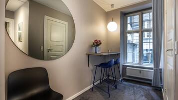 A room with a round mirror, a black chair, a desk with two blue chairs, a radiator, and a window with curtains.