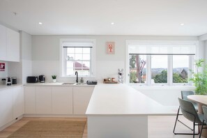 A modern kitchen with a large island, white cabinetry, a window with a view, and a dining area with a table and chairs. - Stunning 3BR with views Walk to Manly (Fairlight)
