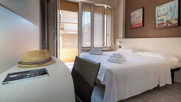 Una camera d'albergo con un letto, una sedia, un tavolo con cappello e libro, e un quadro appeso al muro.