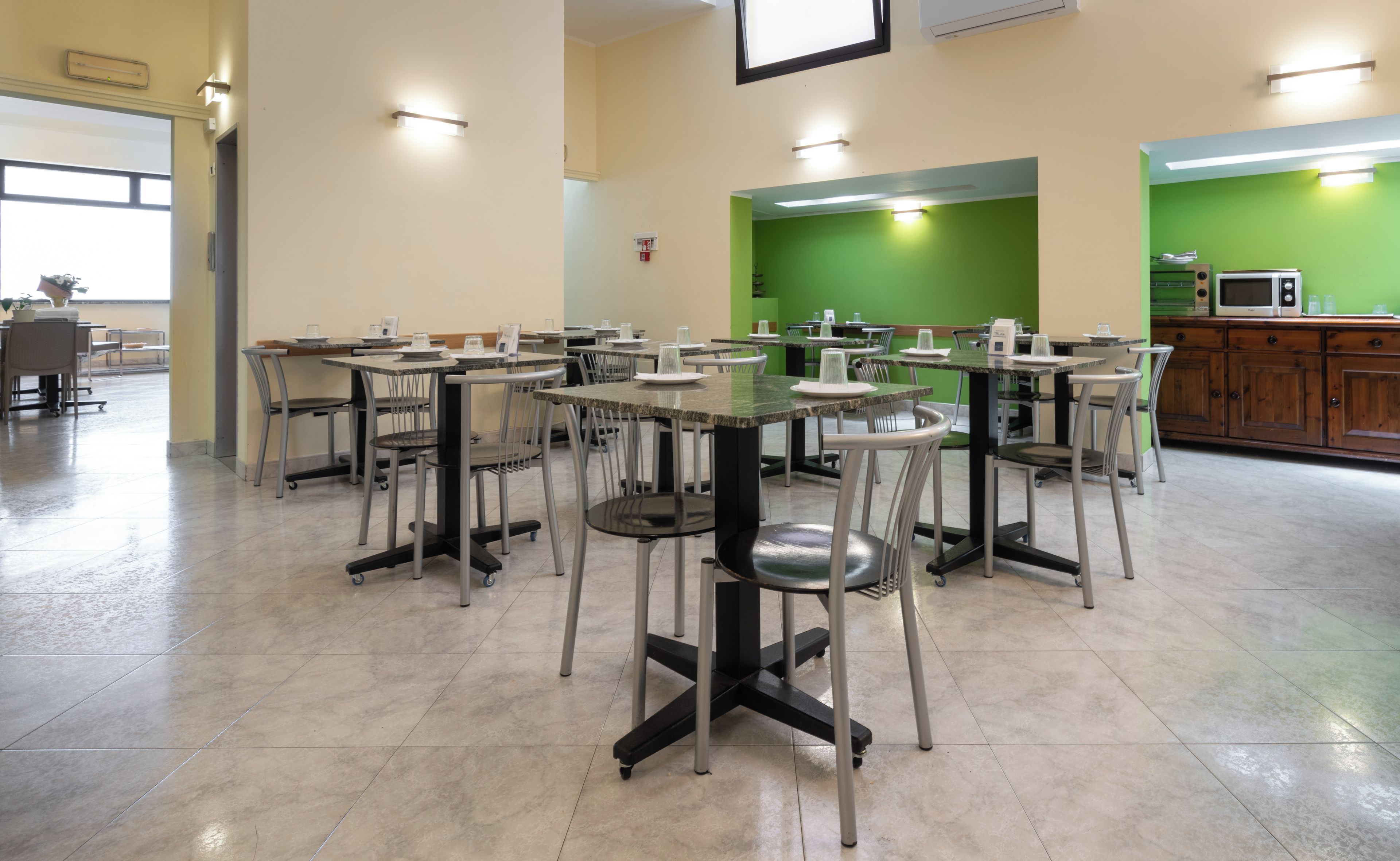 A spacious dining area with multiple tables and chairs, a green accent wall, and a kitchen area with cabinets and appliances.