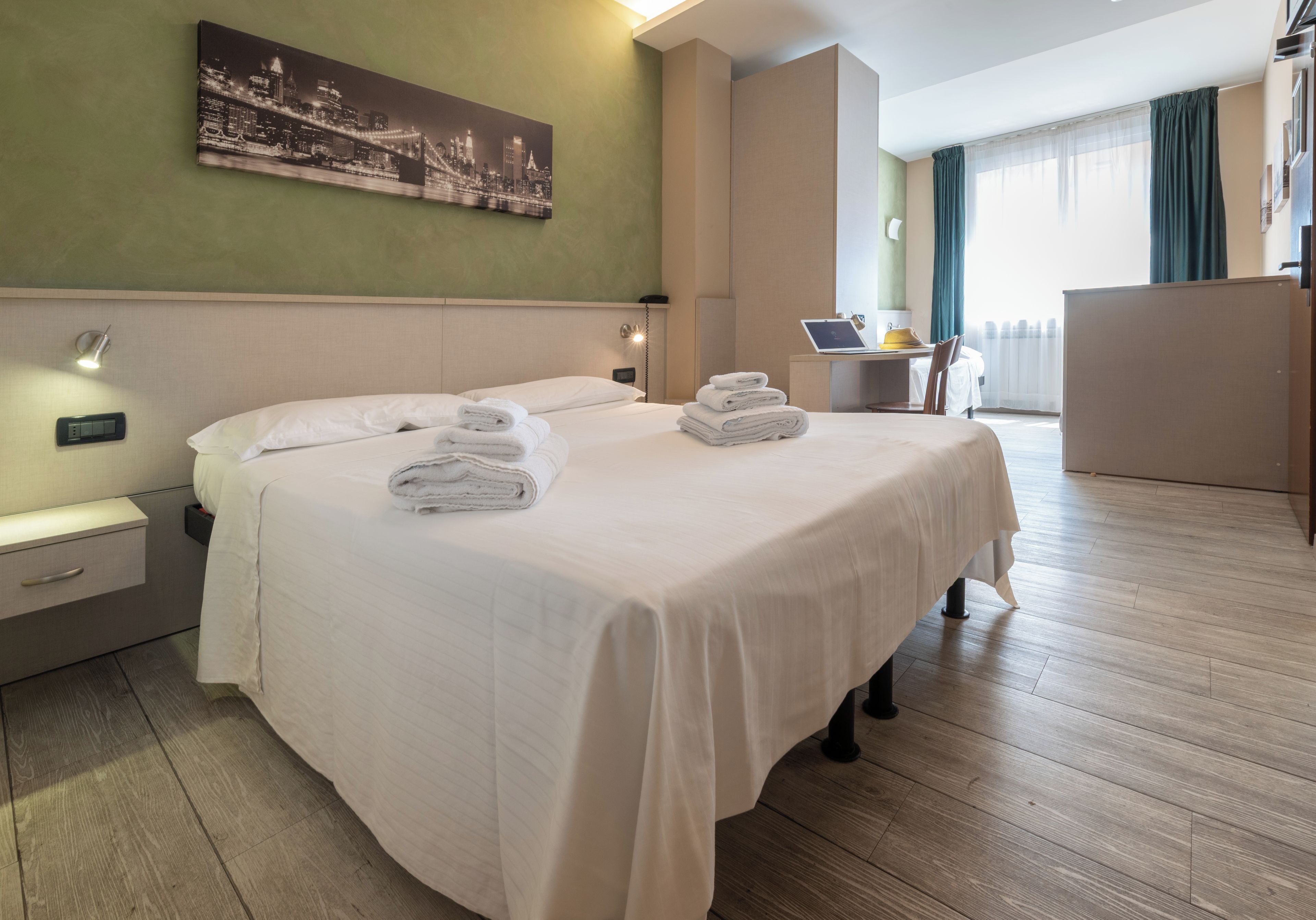 Una habitación de hotel con una cama, un escritorio con una computadora portátil, una silla y un cuadro de un paisaje urbano en la pared.