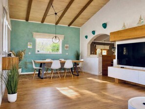 A dining area with a wooden table and chairs, a large window, and a television in the living room. - Finca in Santa Magalida With Wifi (Santa Margalida)