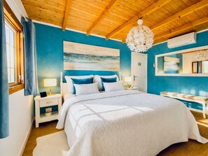 A bedroom with a wooden ceiling, a large bed with white bedding, a nightstand, a bedside table, a wall-mounted air conditioner, and a painting of a beach scene. - Finca in Santa Magalida With Wifi (Santa Margalida)