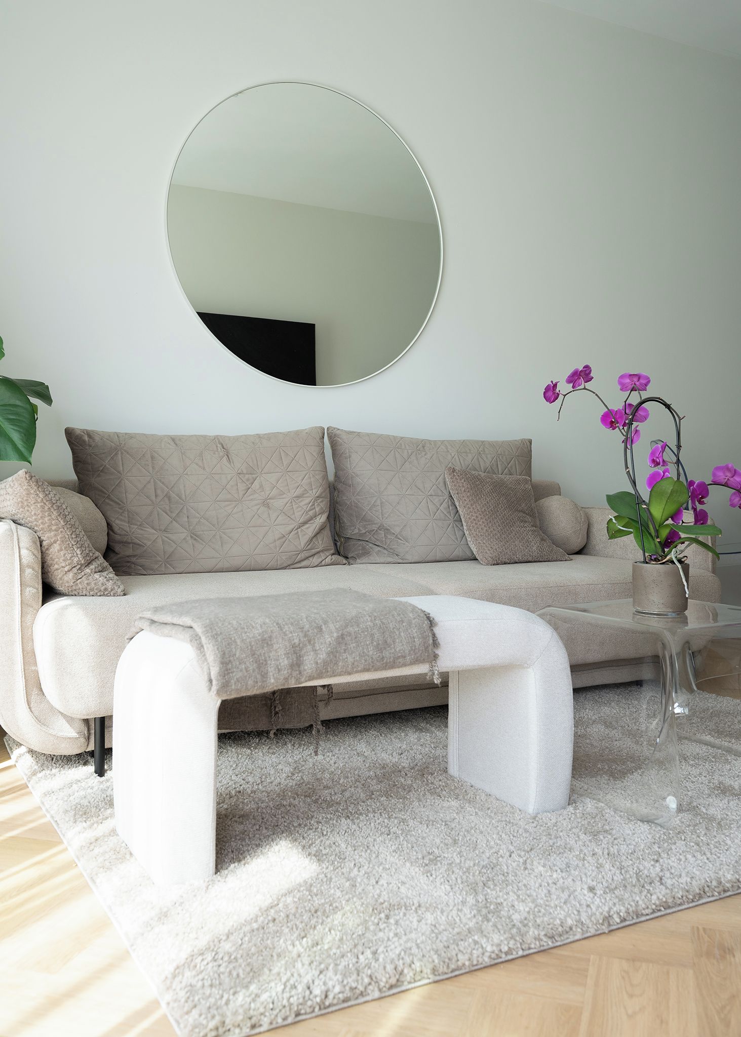 Una sala moderna con un sofá, un espejo redondo, una mesa de centro y una orquídea en maceta.