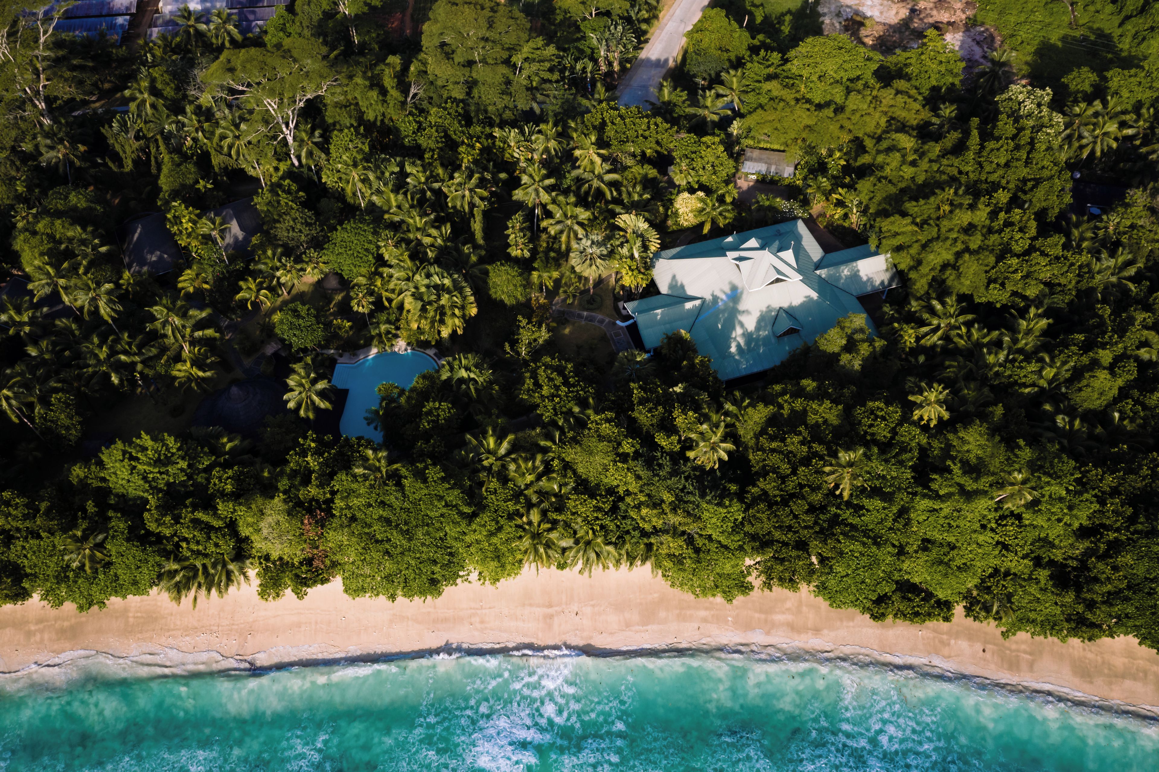 An aerial view of a tropical resort with a beachfront, dense palm trees, and a large building with a unique roof structure.
