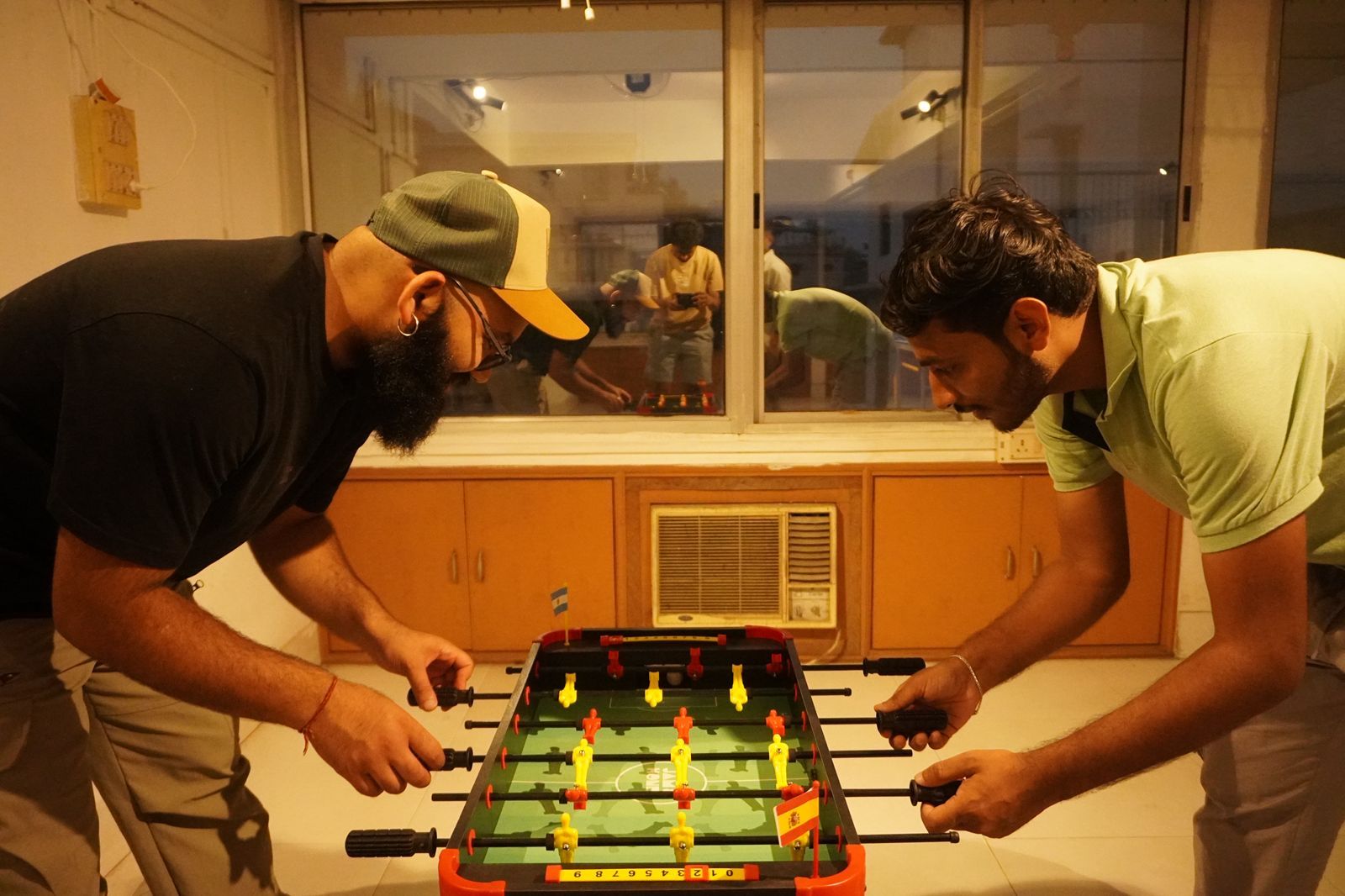 Duas pessoas jogando pebolim em um espaço com armários de madeira e uma janela.
