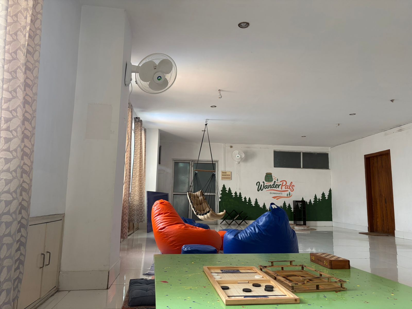 Una habitación con una mesa verde, pufs, una silla colgante y un mural en la pared.