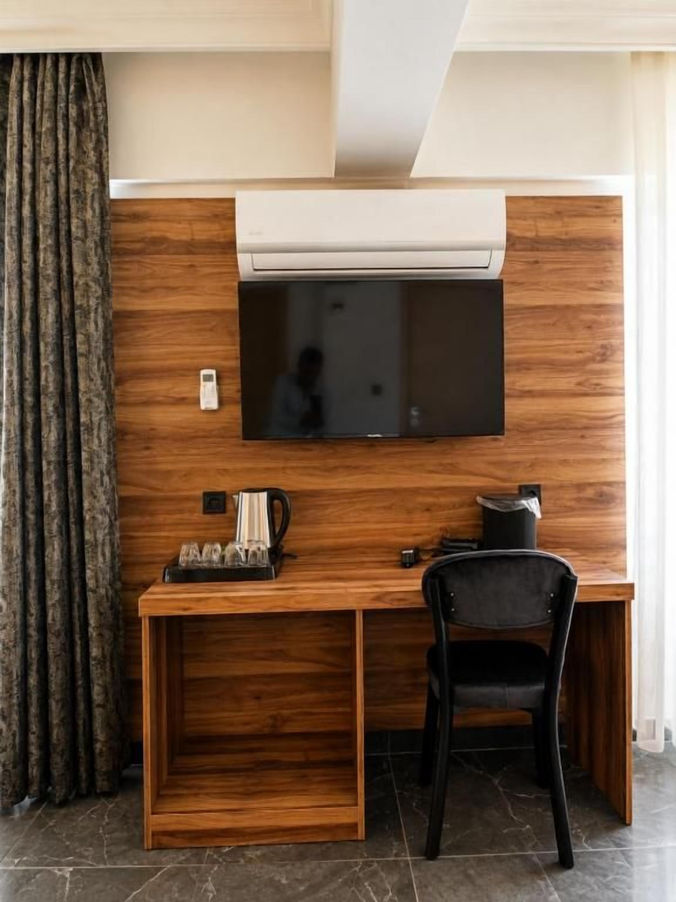 Une pièce de taille modeste, avec un bureau en bois, une chaise noire, un téléviseur à écran plat fixé au mur et une machine à café.