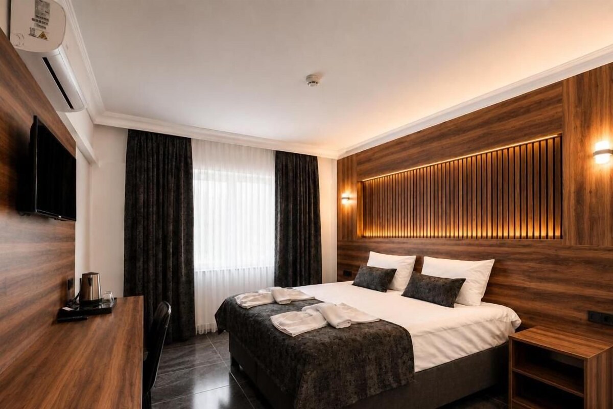 Una habitación de hotel moderna con una cama grande, un televisor de pantalla plana, un panel de pared de madera y una ventana con cortinas.