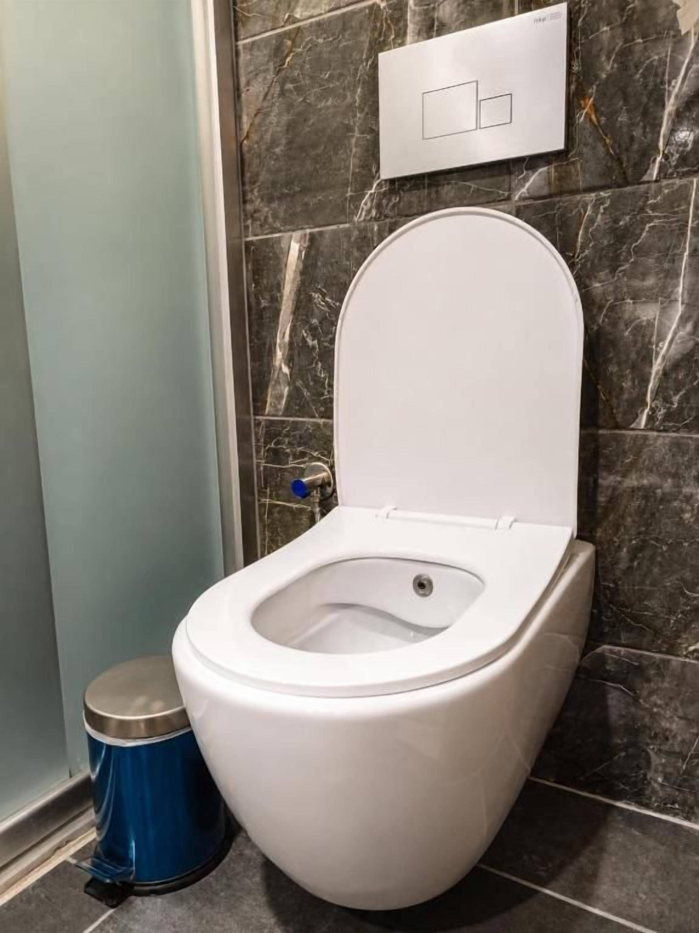 A white toilet with a closed lid, a blue trash bin, and a marble wall with a light switch.