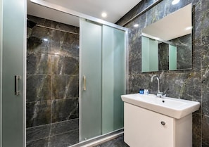 Un baño moderno con cabina de ducha de vidrio, lavabo blanco y pared revestida de mármol.