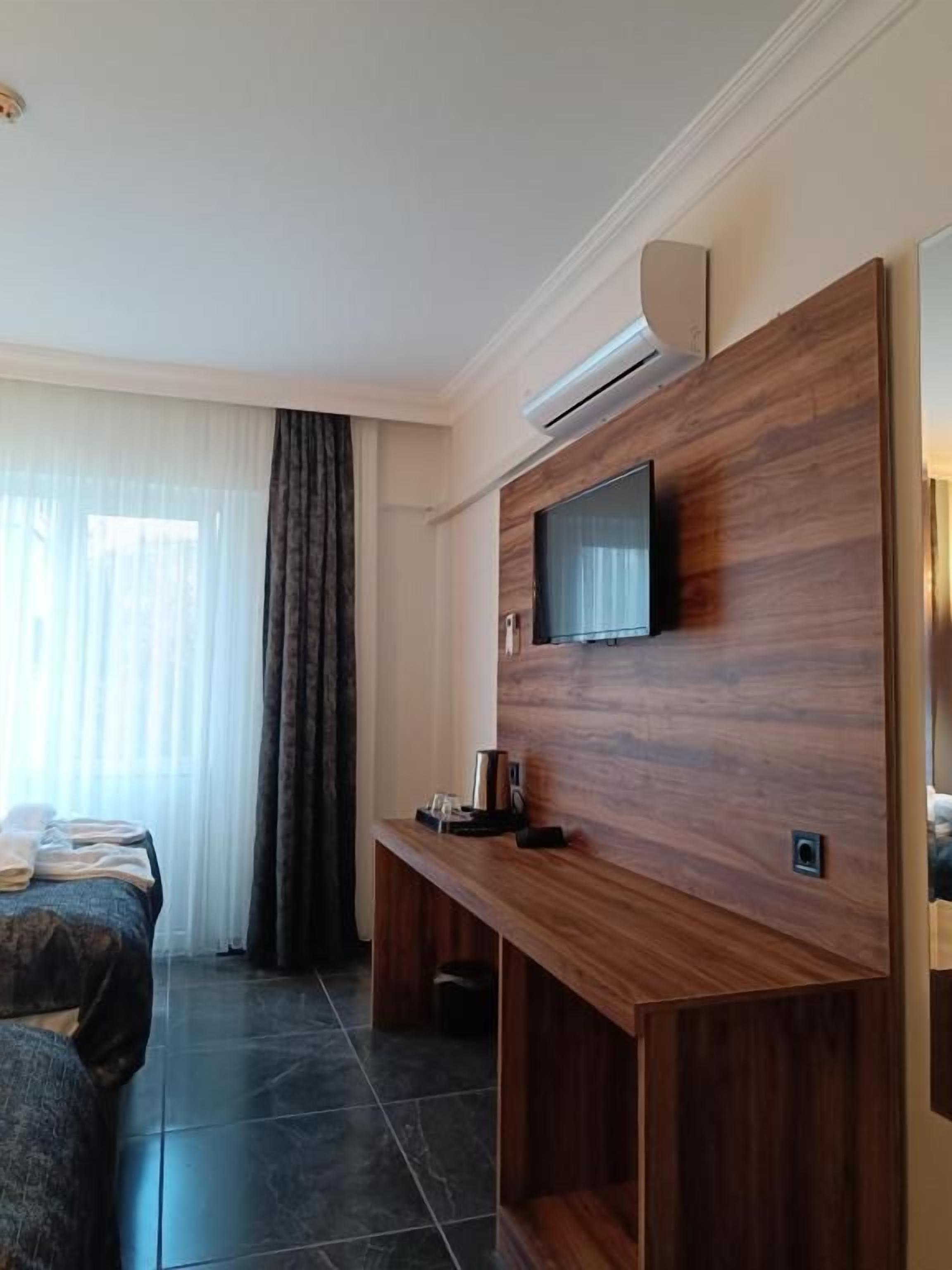 A modern hotel room with a wooden entertainment unit, a flat-screen TV, a bed, and a large window with curtains.