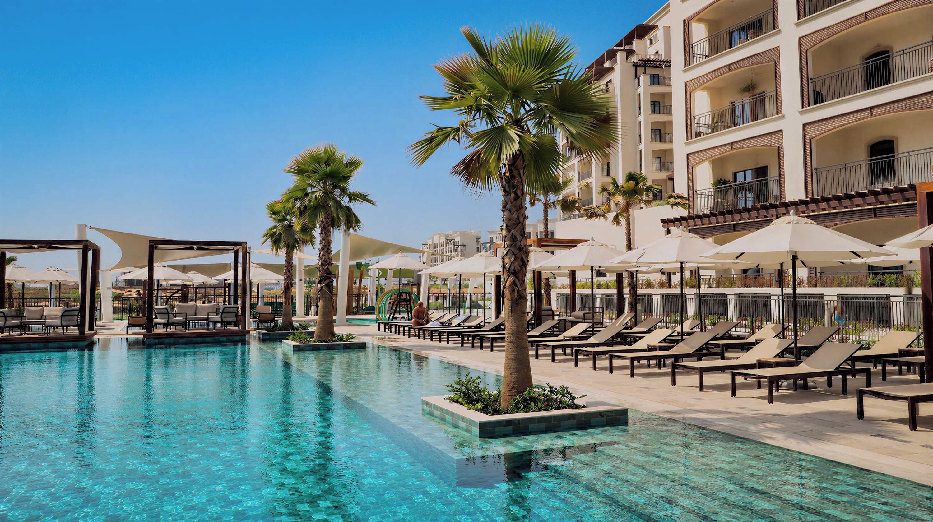 A pool area with lounge chairs and umbrellas, surrounded by palm trees and a multi-story building.