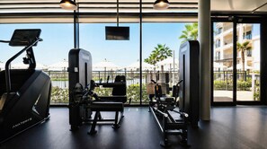 A gym with cardio machines, weight benches, and a view of a pool and palm trees through large windows. - Vanilla Homes Premium Studio Yas Golf Collection (Abu Dhabi)