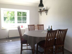 A dining area with a round table, six wooden chairs, a window with a view of greenery, a radiator, and a shelf with a lamp and items. - Crildumersiel Farm (Wangerland)