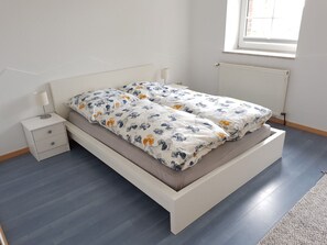 A bed with floral bedding, a bedside table with a lamp, and a window with blinds. - Crildumersiel Farm (Wangerland)