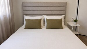 Uma cama com cabeceira, duas almofadas, mesa de cabeceira com uma planta e cortina.