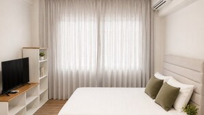 Quarto com cama, televisão em móvel, mesa de cabeceira e janela com cortinas.