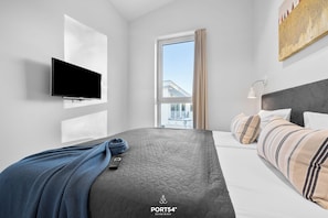 A modern hotel room with a large bed, a flat-screen TV mounted on the wall, a window with curtains, and a city view. - Exclusive Penthouse Apartment with Sauna and Balcony near Priwall Beach (Lübeck)