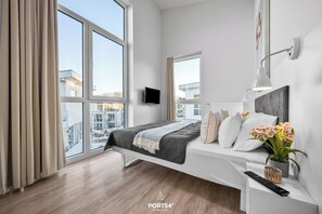 Une chambre moderne avec une grande fenêtre, un lit recouvert d’une couverture sombre, une table de chevet avec une lampe et une télécommande.