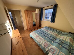 A bedroom with a wooden wardrobe, a bed with a patterned bedspread, a hanging light fixture, and a window with curtains. - Dünenzauber – Your Relaxing Getaway at Schönberger Strand (Schönberg (Holstein))
