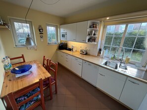 A kitchen with white cabinets, a wooden dining table, and a window with a view of greenery. - Dünenzauber – Your Relaxing Getaway at Schönberger Strand (Schönberg (Holstein))