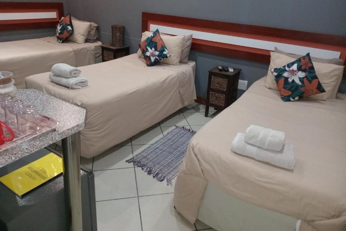 Een hotelkamer met drie bedden, een klein tafeltje met een fles en een geel kaartje, en een afstandsbediening op een nachtkastje.