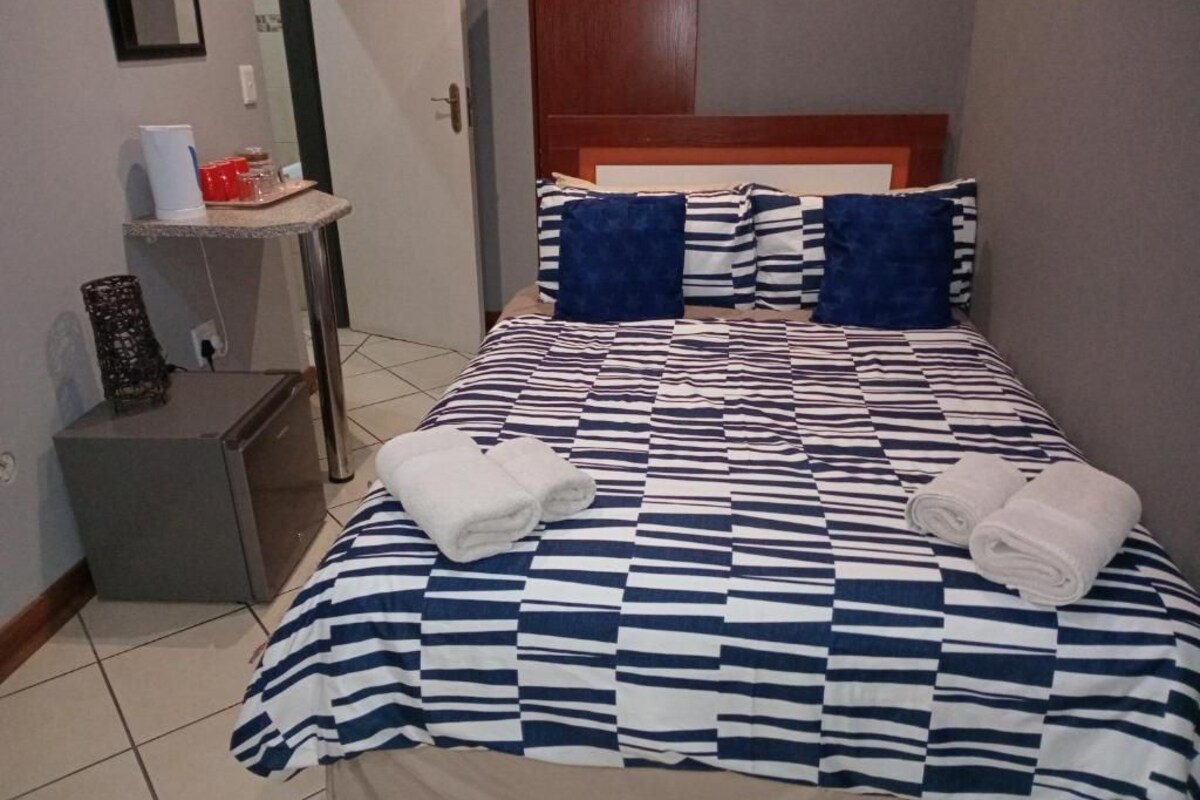 Hotellihuone, jossa on ruutukuviollinen peitto sängyllä, kaksi tyynyä, yöpöytä lampun kanssa ja pieni kaappi.