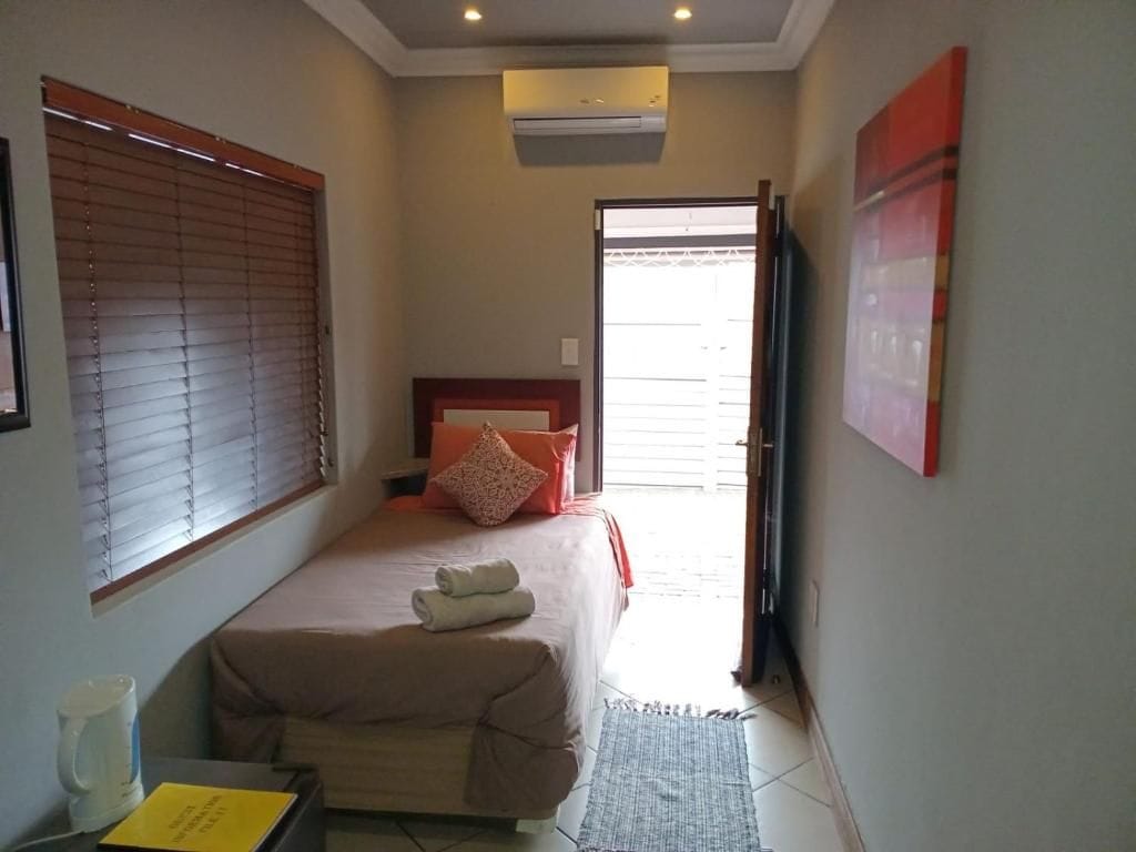 A bedroom with a bed, a bedside table, a window with blinds, an air conditioning unit, and a door leading to another room.
