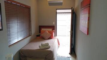 A bedroom with a bed, a bedside table, a window with blinds, an air conditioning unit, and a door leading to another room.