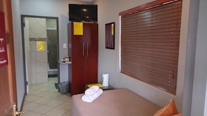 A hotel room with a bed, a television, a window with blinds, a wardrobe, and a reception area visible through an open door. - Grand Central Guesthouse (Rustenburg)