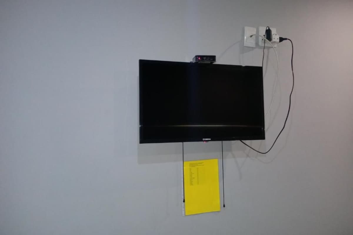 Tasainen seinä, jossa on kiinnitettynä litteä televisio, ja sen alla on näkyvissä virtajohto sekä keltainen muistilappu.