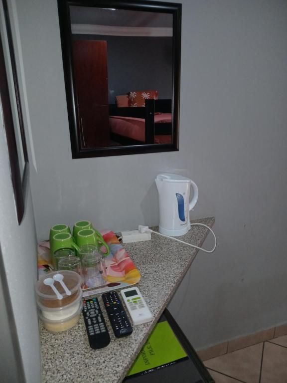 Habitación de hotel con cama, espejo, hervidor eléctrico, cafetera, bandeja con tazas y vasos, y control remoto.