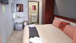A hotel room with a bed, a desk, a mirror, and a television. - Grand Central Guesthouse (Rustenburg)