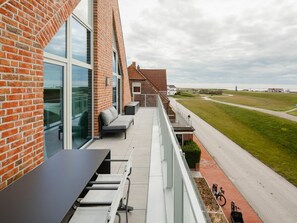 A balcony with a seating area, a table, and a view of a grassy area and a distant structure. - Meer.loft.juist.9 (Juist)