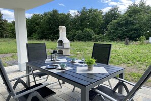 Eine Terrasse im Freien mit Esstisch und Stühlen, im Hintergrund ein Steinofen.