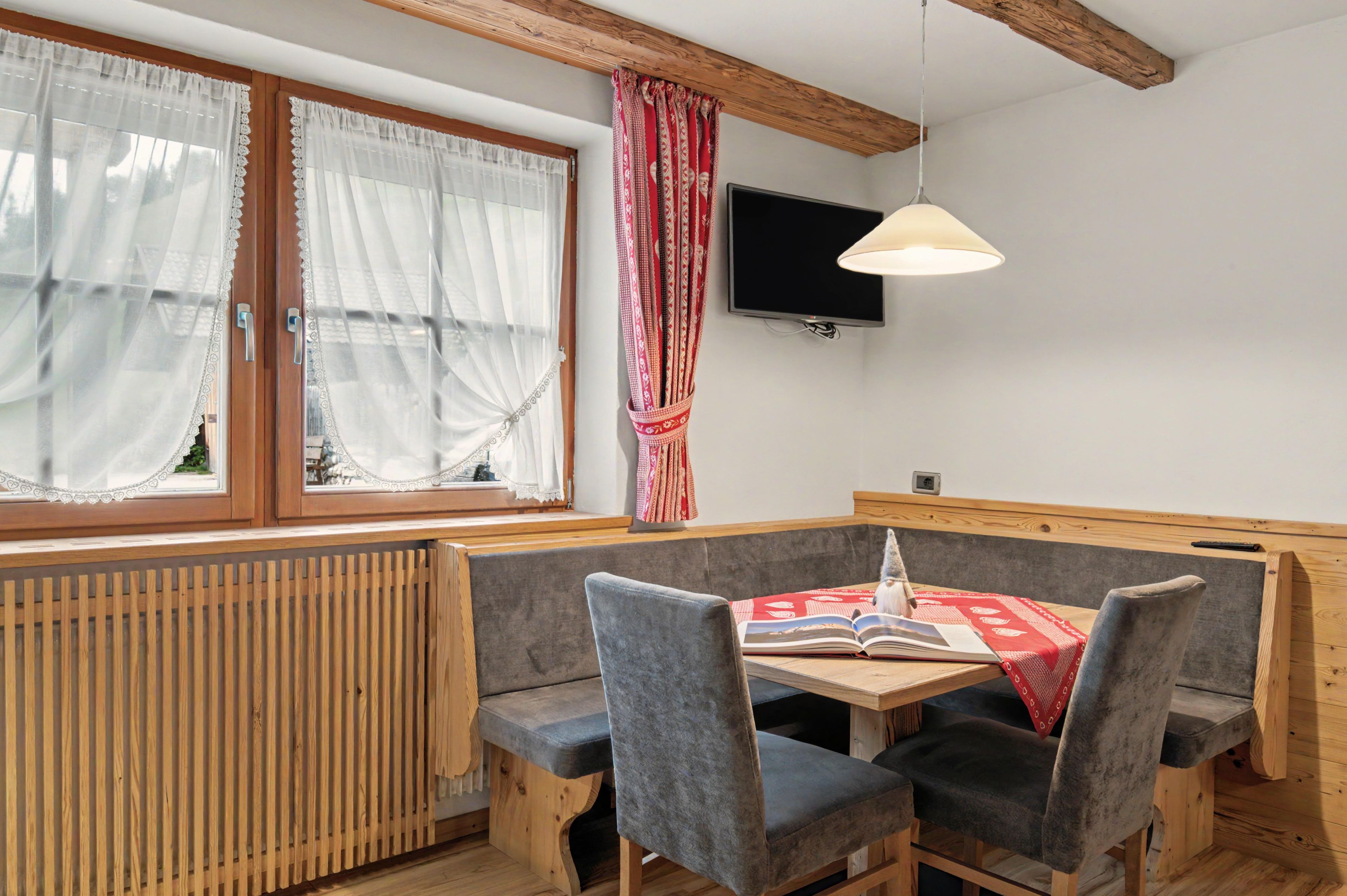 A cozy room with a wooden bench, a table with a red patterned tablecloth, and a television mounted on the wall.