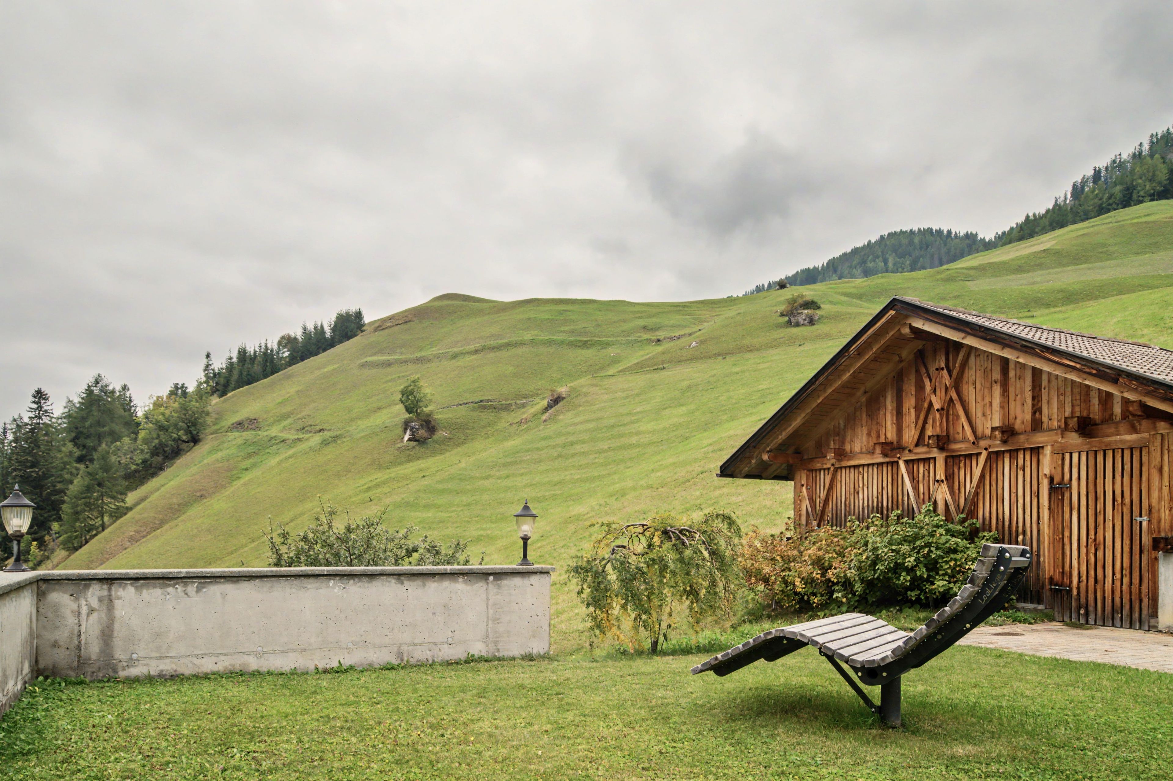 A wooden cabin with a sloped roof, a concrete wall, and a metal bench in a grassy area with a hill in the background.