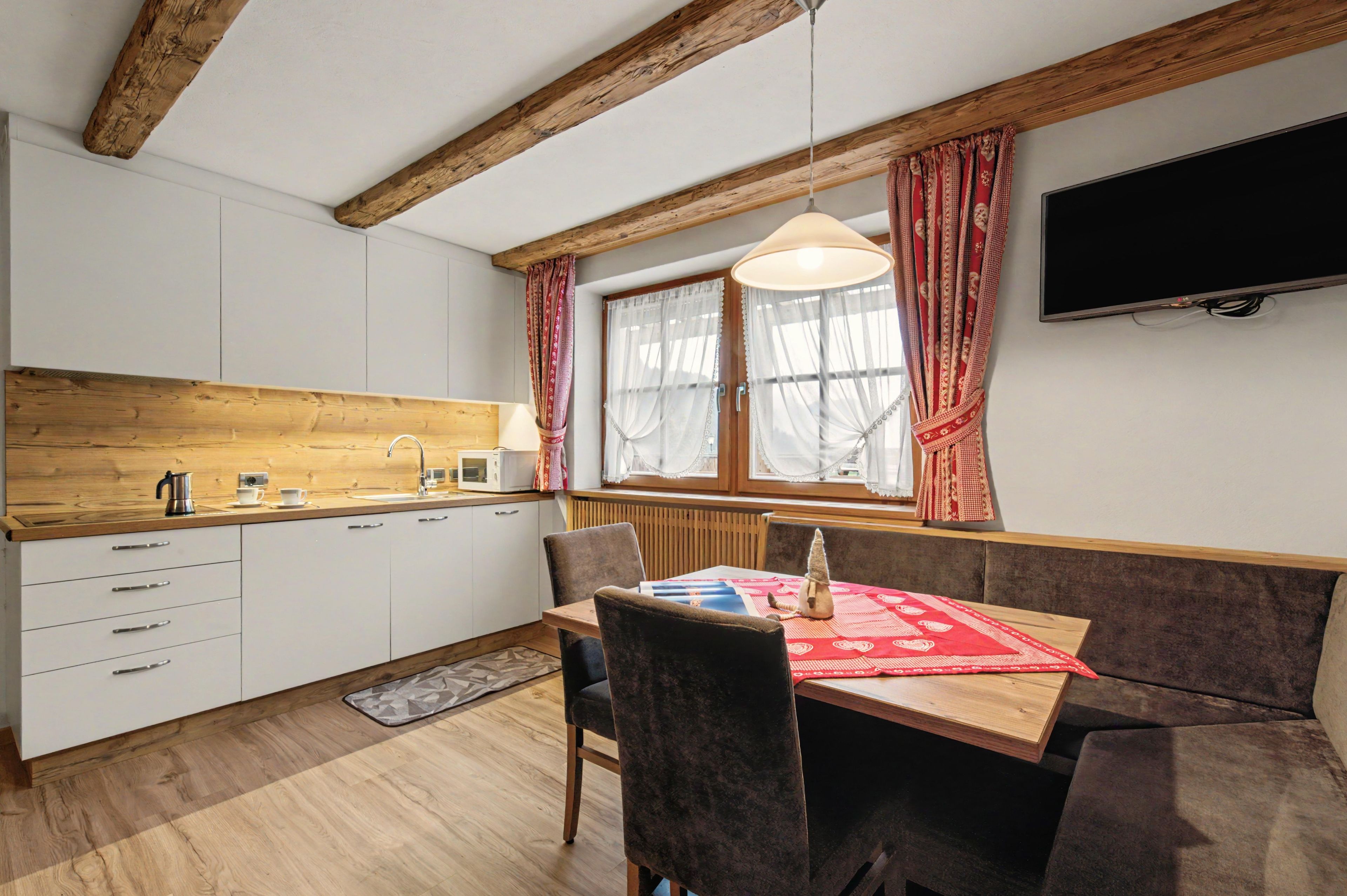 A compact kitchen with wooden beams, a dining area with a table and chairs, and a TV mounted on the wall.