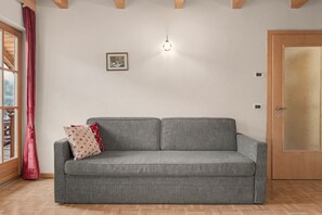 A grey sofa with two pillows, a wooden door with a frosted glass panel, a small framed picture on the wall, and a window with a red curtain. - Apartment 'Wirthof  4' with Mountain View, Shared Terrace and Wi-Fi (La Valle)