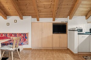 A compact kitchen with wooden cabinets, a built-in TV, and a dining area with a patterned tablecloth. - Apartment 'Wirthof  4' with Mountain View, Shared Terrace and Wi-Fi (La Valle)