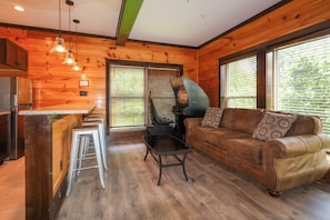 A cozy cabin interior with a kitchenette, a seating area with a sofa and a coffee table, and a large window with blinds. - Private indoor pool, driving sim games, pool table, Smoky Mountain views! (Gatlinburg)